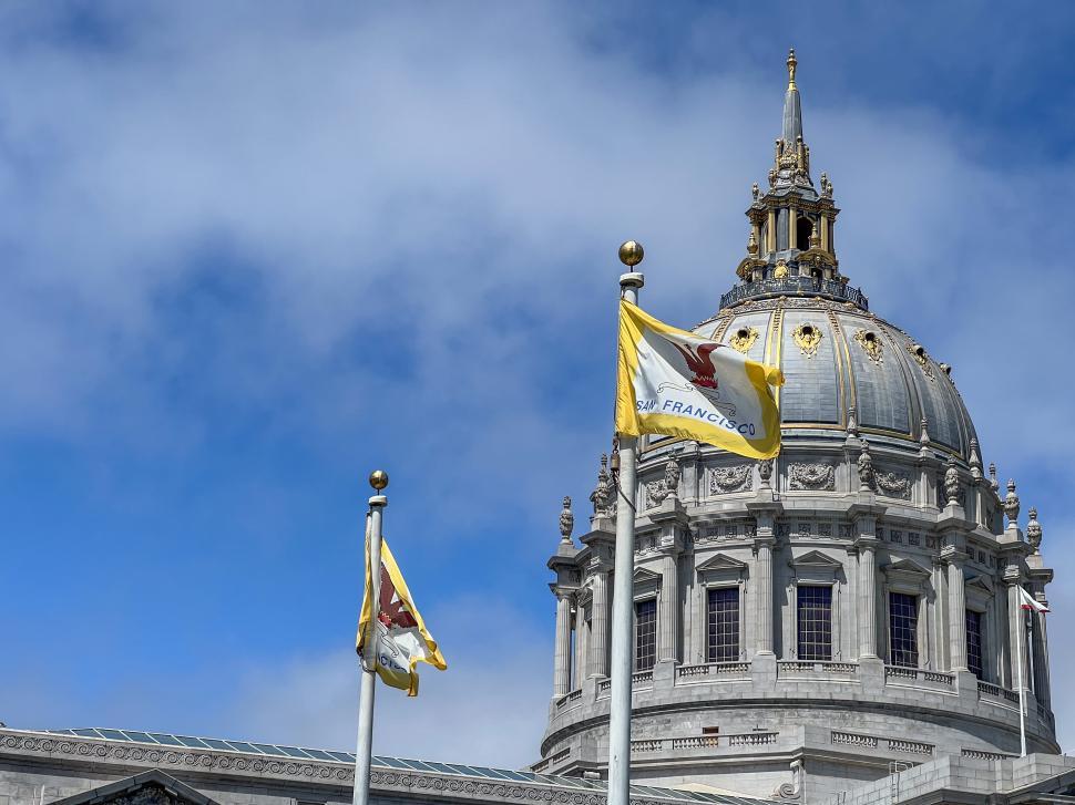 Free Image of Historic San Francisco city hall dome under a partly cloudy sky with flags waving 