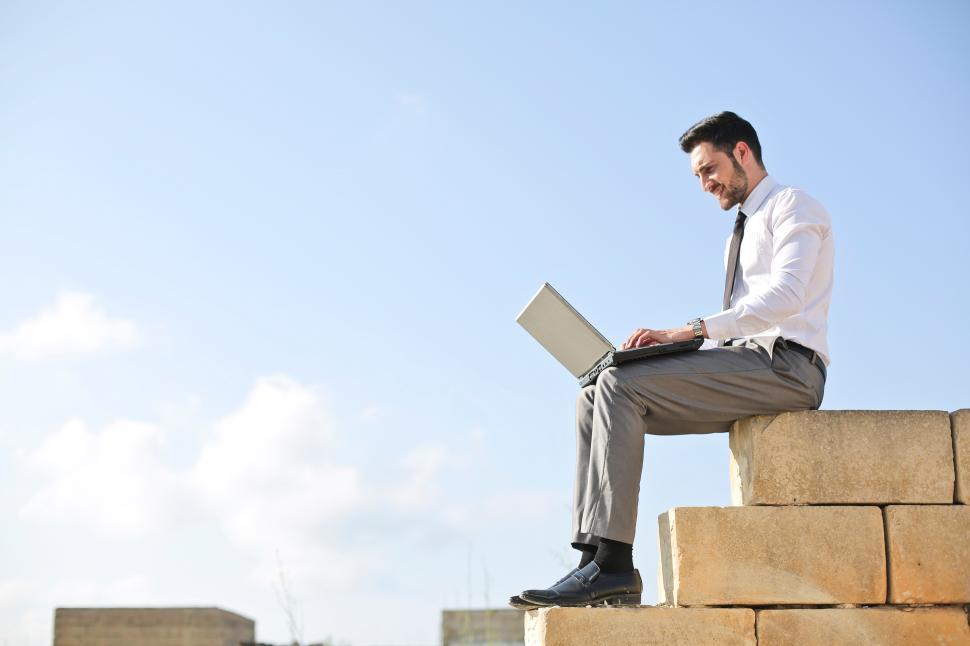 Free Image of Businessman working on laptop while sitting on a stone wall outdoors 