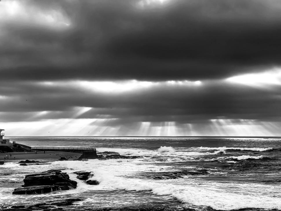 Free Image of Black and white image of waves on rocky shore as stormy clouds part at sunset near ocean 