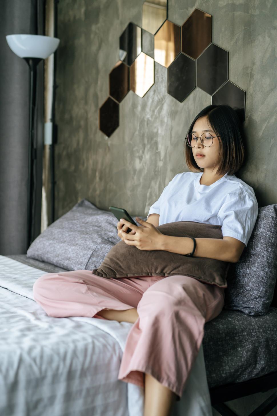 Free Image of Woman wearing a white shirt lying on bed and playing smartphone. 