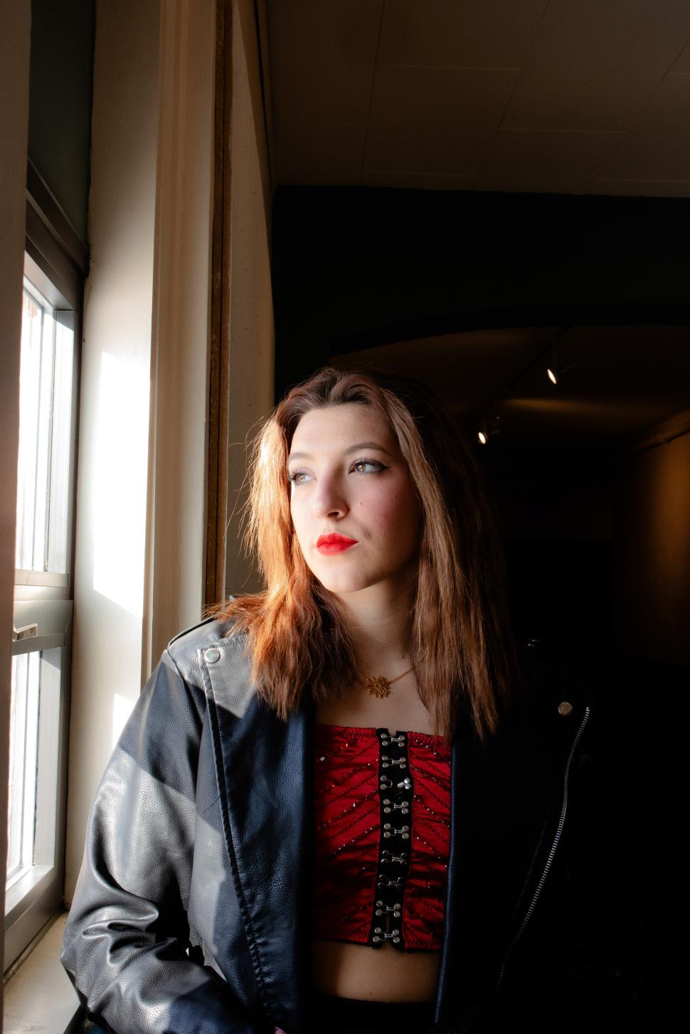 Free Image of Woman in stylish outfit gazes out a window in a dimly lit room 