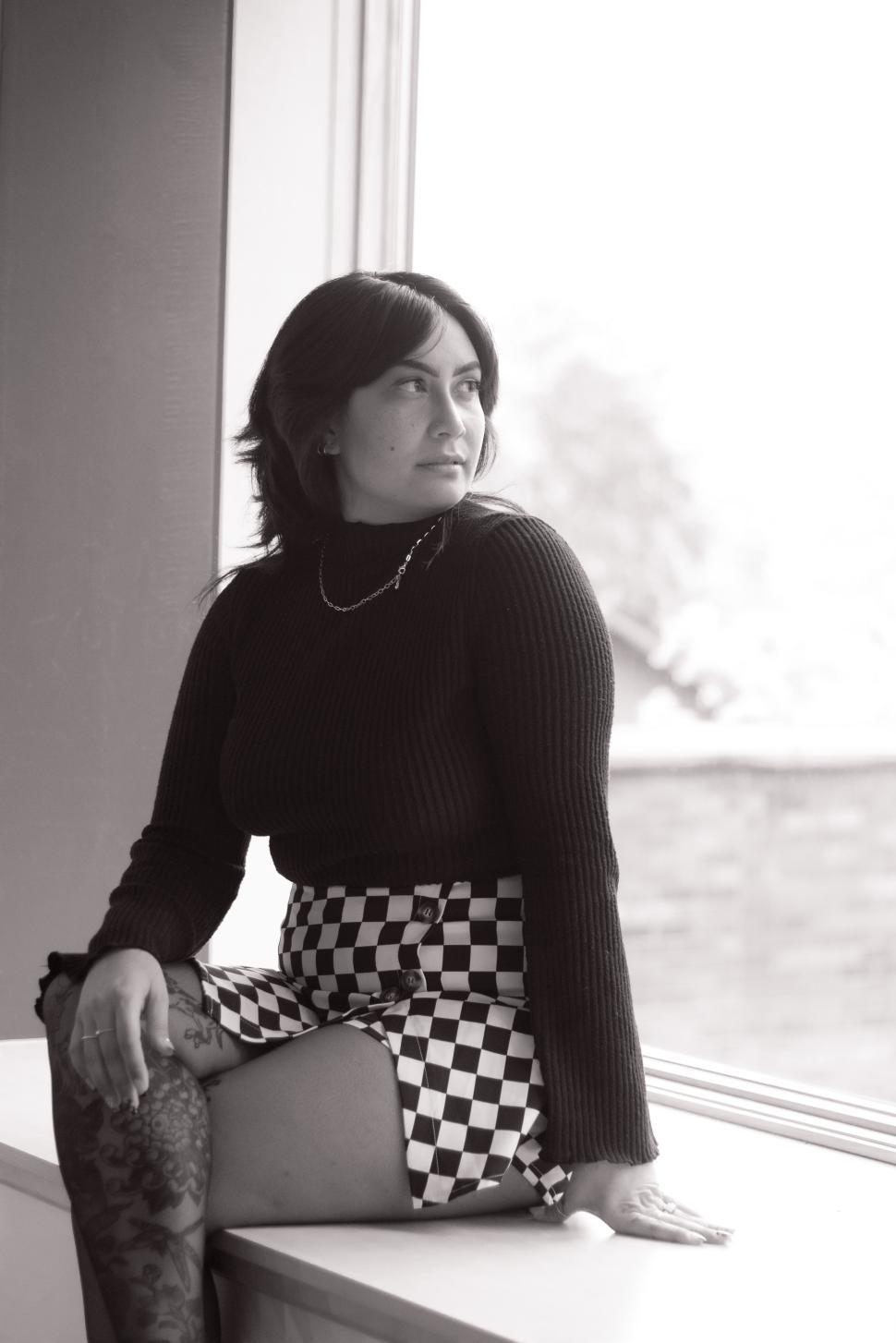 Free Image of Young woman sitting by a window in a black sweater and checkered skirt 