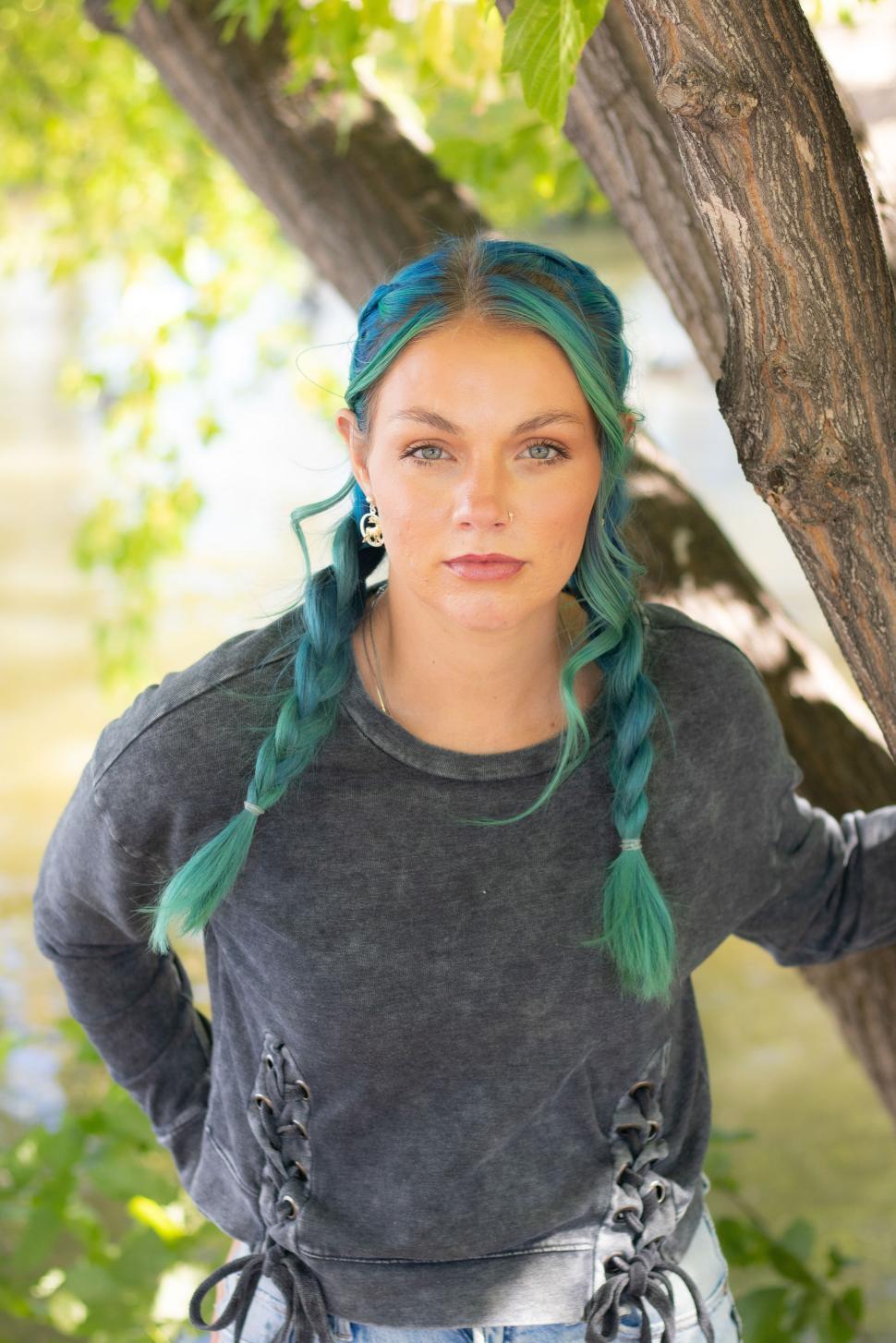 Free Image of Woman with turquoise hair poses by a river surrounded by greenery during daytime 