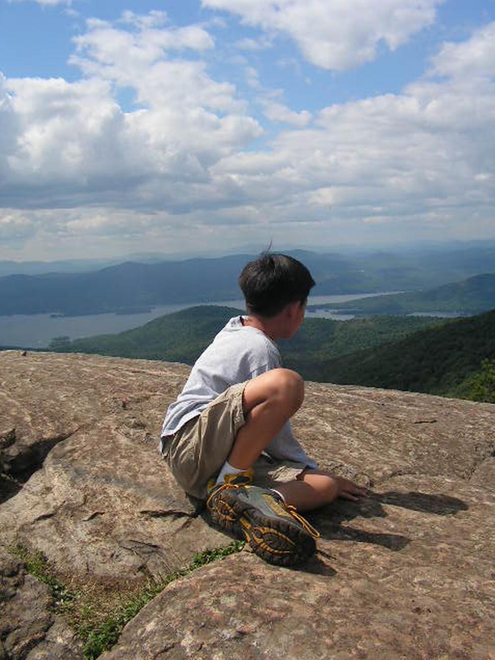 Free Image of This image shows a young hiker resting after a hike to a clearing on a mountain top taking in the view of surrounding mountains and lakes. 