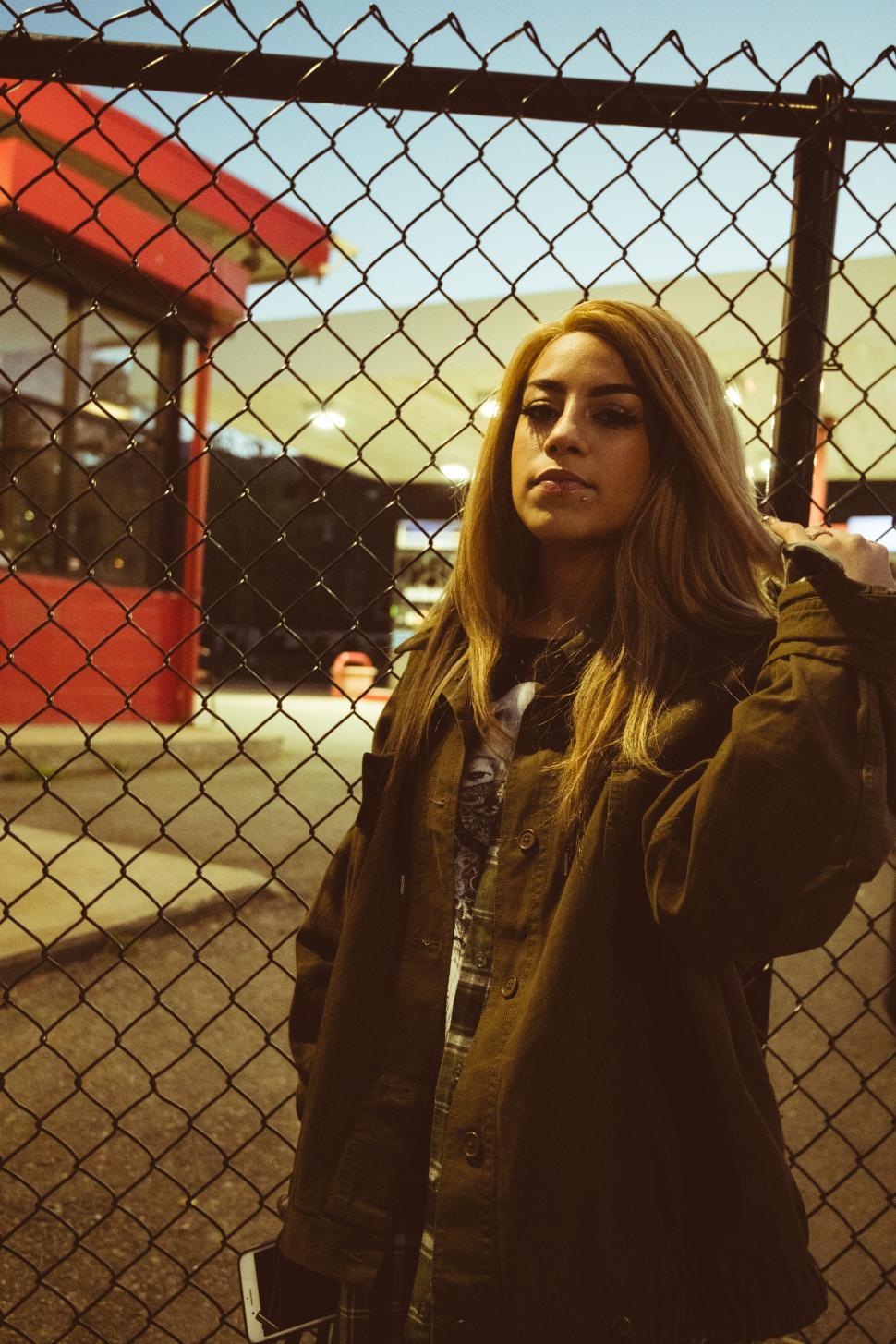 Free Image of Woman posing near a fence in an urban setting at dusk 