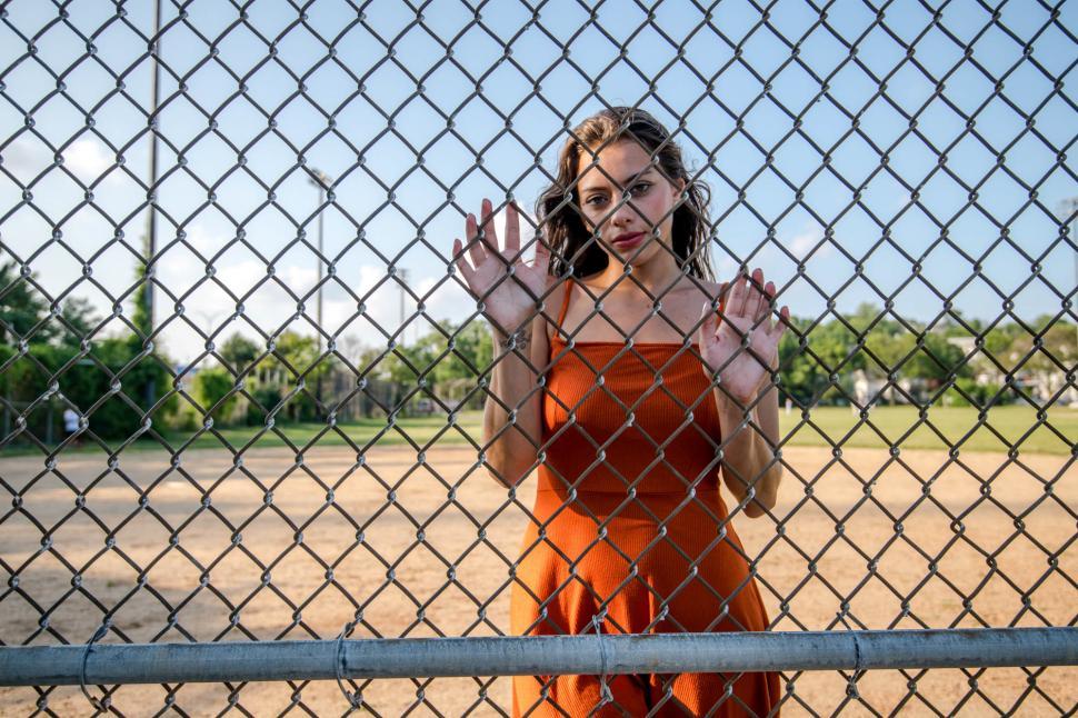 Free Image of Woman in orange dress stands by chain-link fence 