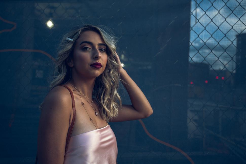 Free Image of Woman with wavy hair poses by a chain link fence at sunset 