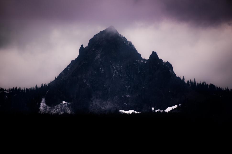 Free Image of Dramatic mountain peak under cloudy sky with snow patches at its base 