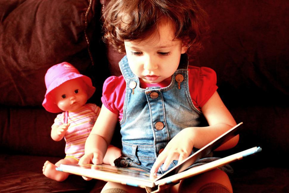 Free Image of A child pretends to be reading a book accompanied by a doll 