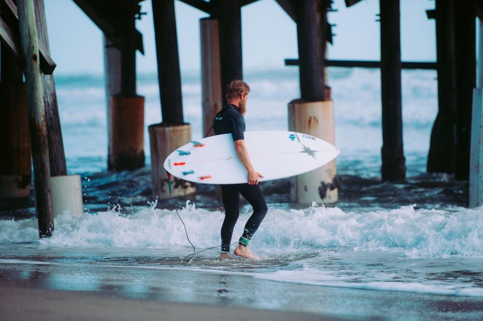 Free Image of Man in Wet Suit Carrying Surfboard Under a Pier 